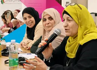 A Jordanian woman with glasses speaks into a microphone as other women listen closely
